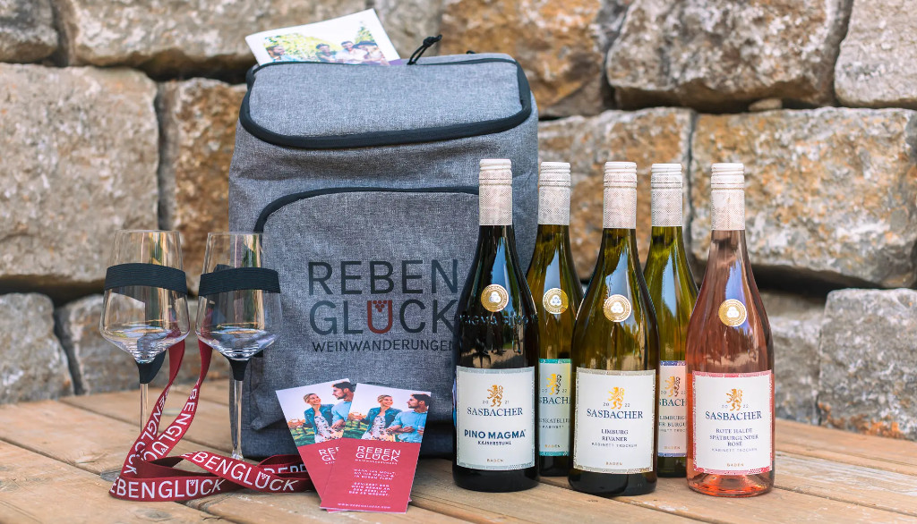 Fünf Flaschen Sasbacher Wein, zwei Weingläser und der Rucksach von Rebenglück stehen auf einem Holztisch vor einer schönen Steinwand